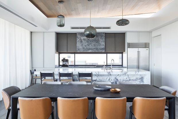 A refined open-plan kitchen and dining area with a large dark dining table in the foreground, tan upholstered dining chairs, and a marble-look kitchen island and benchtops beyond. The kitchen features white cabinetry, dark timber-toned overhead cabinetry, a stainless-steel refrigerator, integrated appliances, a wide horizontal window, and a timber-lined ceiling with contemporary pendant lighting.