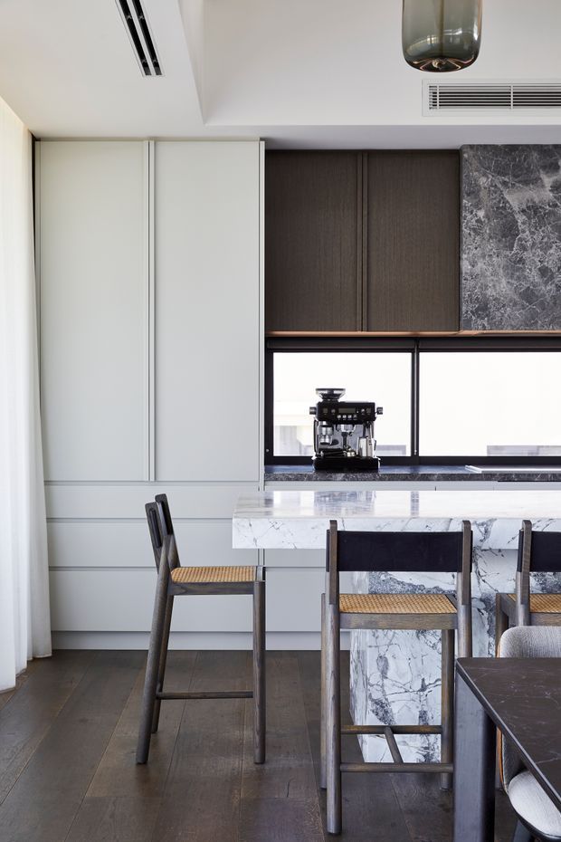 A refined contemporary kitchen viewed from the dining area, featuring full-height pale cabinetry, dark timber overhead joinery, a dramatic grey veined marble splashback and island, dark timber flooring, and a compact coffee station beneath the window. Woven-seat timber bar stools and dining chairs add warmth to the otherwise restrained palette.