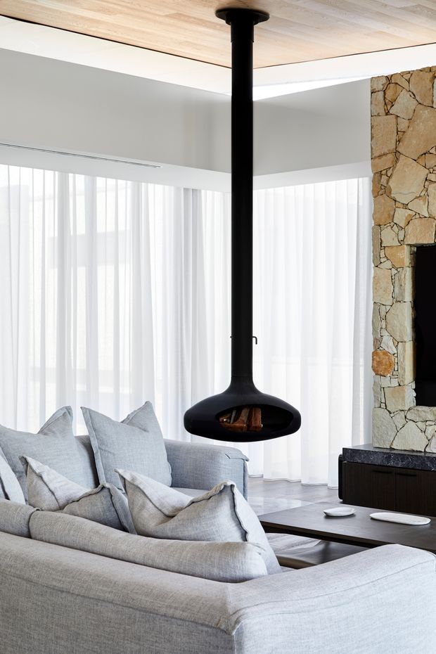 Bright contemporary living room with a large pale upholstered sectional sofa, sheer full-height curtains, a suspended black fireplace, light timber ceiling, and a rugged natural-stone fireplace wall with dark built-in cabinetry. The room has abundant filtered daylight and a refined neutral palette.