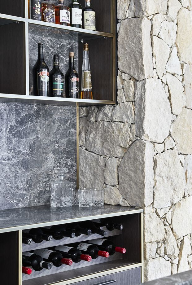 Close-up of a built-in home bar and wine storage feature. Dark timber cabinetry with open bottle shelving, a dark veined stone benchtop and splashback, glassware, spirits, and horizontal wine racks are set against a prominent rough-faced light natural-stone wall.
