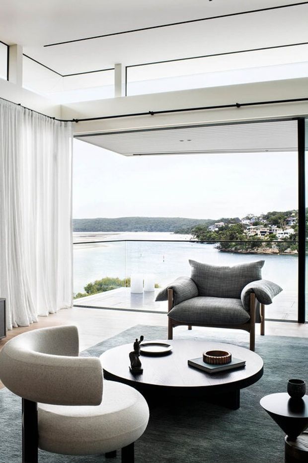A refined contemporary living room with floor-to-ceiling glazing overlooking a calm waterfront inlet. The room features sheer white curtains, a dark-framed operable glazed opening, pale flooring, a large muted rug, sculptural upholstered lounge chairs, and a low round dark coffee table with restrained decor. The space is bright, open, and strongly connected to the exterior view.