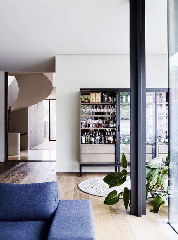 A refined contemporary living area with a deep blue sectional sofa, pale timber flooring, and expansive black-framed glazing. A freestanding built-in-style drinks and wine cabinet occupies the central wall, while a sculptural curved staircase and open circulation space are visible beyond. Indoor plants and a textured round rug add softness to the clean, light-filled interior.