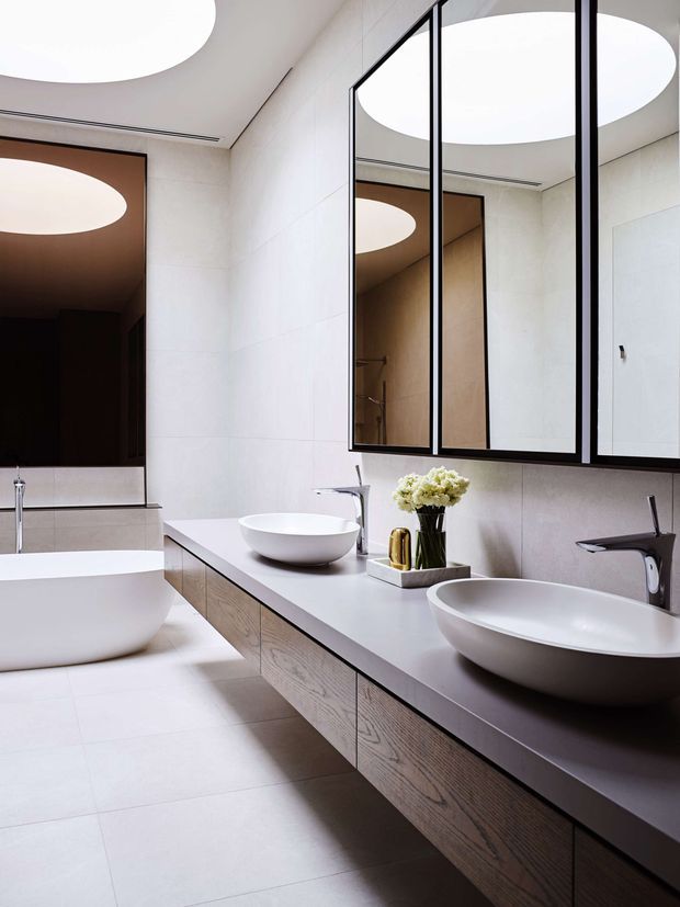 Contemporary bathroom featuring a long floating timber vanity with two white vessel basins, wall-mounted dark faucets, large black-framed mirrored cabinets, pale tiled walls and floor, and dramatic circular ceiling lights. A freestanding or partially visible bathtub and a dark shower or adjoining zone appear at left.