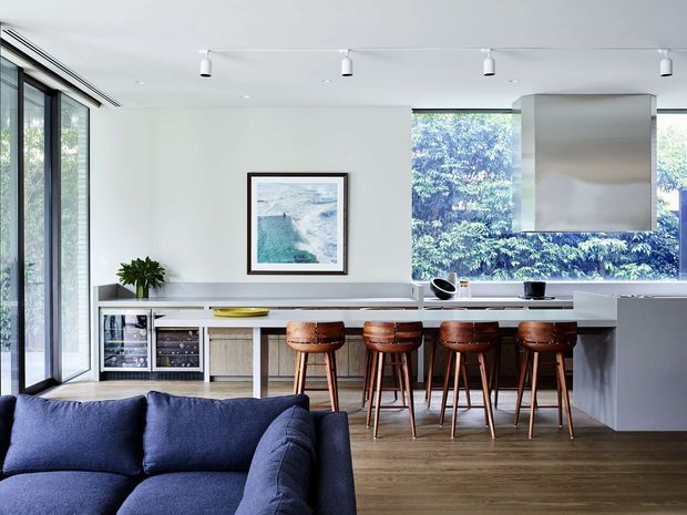 A bright, open-plan contemporary kitchen and living area. The kitchen features a long light-toned island or breakfast bar with four timber bar stools, integrated wine storage, light wood cabinetry, and a large stainless-steel rangehood. Floor-to-ceiling glazing provides views of greenery, while a deep navy sectional sofa occupies the foreground. White walls, timber flooring, streamlined track lighting, and minimal furnishings create a clean, refined interior.