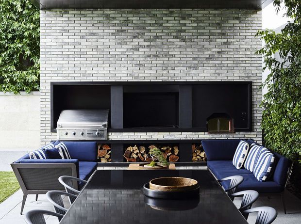 A contemporary outdoor entertaining area with a built-in black outdoor kitchen and barbecue set into a light grey brick wall. A long recessed wood-storage niche runs beneath the cooking appliances, while a dark outdoor dining table and blue cushioned seating occupy the foreground. Dense greenery frames both sides of the compact courtyard-like space.