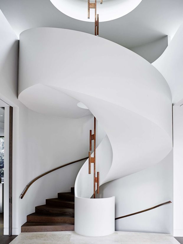 A sculptural interior stairway dominated by a sweeping, curved white plaster or painted form that wraps around a dark timber spiral staircase. Slim brown metal or timber handrails follow the curved walls, with vertical warm-toned balustrade elements rising toward a circular ceiling opening and pendant light. The space is bright, minimalist, and highly architectural, with pale stone flooring and smooth white surfaces.