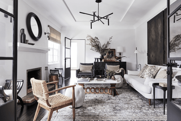 A refined living room with white walls, a traditional fireplace, black-framed glass doors and partitions, a white sofa, woven armchair, patterned area rug, sculptural coffee table, dark sideboard, and contemporary linear pendant lighting. The room blends classic architectural elements with modern and eclectic furnishings.