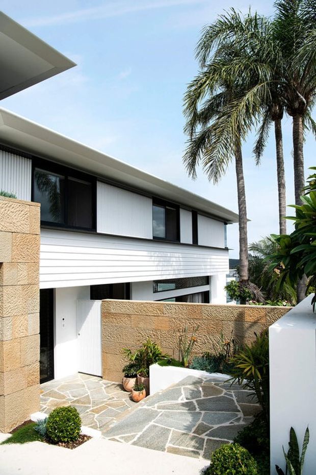 Exterior view of a contemporary multi-level residence with crisp white horizontal cladding, dark-framed glazing, projecting flat roof planes, and textured tan masonry walls. A patterned stone-paved entry courtyard is landscaped with tropical palms and planting.