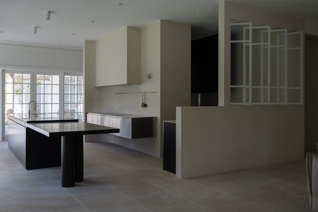 A refined open-plan kitchen and dining area with a large black island, integrated pale cabinetry, tiled flooring, and a partially screened stair or hallway zone. White-framed glazed doors and windows bring in natural light, while the restrained palette and clean lines create a contemporary, minimalist feel.