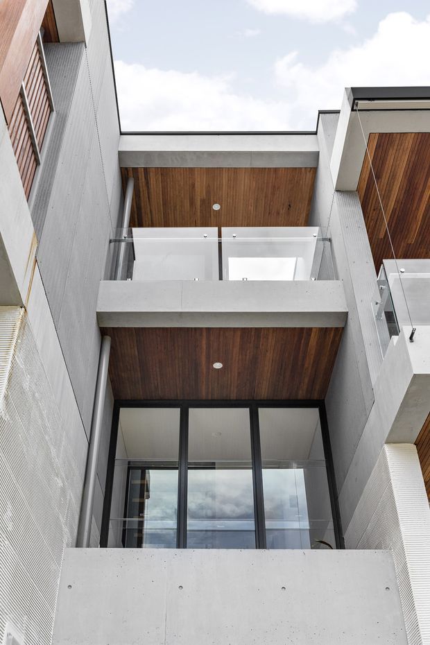 Upward-looking view of a contemporary multi-level building courtyard or facade, featuring exposed concrete balconies, clear glass balustrades, dark aluminium-framed glazing, warm timber soffits, and vertically ribbed light-coloured wall cladding. The composition emphasizes layered balconies, an open central void, and clean geometric lines against the sky.