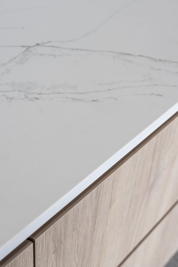 Close-up detail of a pale grey, marble-look benchtop with subtle veining, set above light natural timber-grain cabinetry with clean, contemporary drawer fronts.