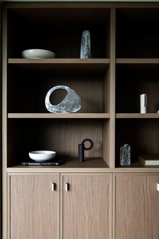 Close-up view of a built-in dark timber display cabinet and lower storage joinery. Open shelves contain minimalist ceramic and stone decorative objects, books, and a small black sculptural lamp or vessel; the lower section has light natural timber-grain doors with simple metal pulls. The composition is warm, refined, and contemporary.