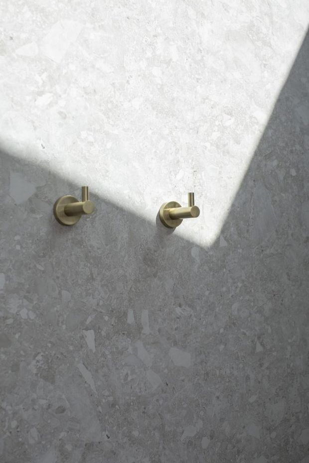 Close-up of two minimalist brushed brass wall hooks mounted on a grey, naturally mottled stone wall. Strong diagonal sunlight and shadow create a high-contrast, refined bathroom-like architectural detail.