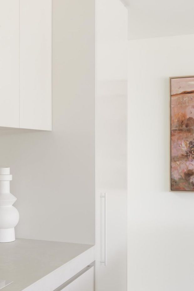 Close-up interior view of a minimalist white built-in joinery installation, with tall flat-panel cabinetry, a pale stone or composite benchtop, integrated vertical handles, and a small white sculptural vase. A framed abstract artwork adds a muted blush and brown accent on the adjacent white wall.