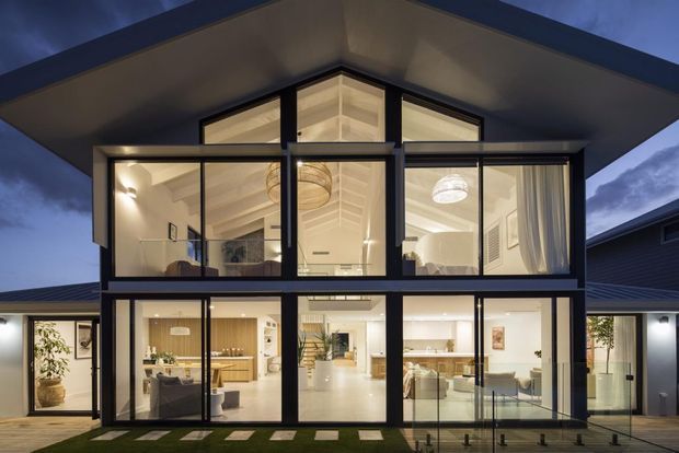 Twilight exterior view of a contemporary two-storey residence with a dramatic steeply angled roof, extensive black-framed floor-to-ceiling glazing, and a bright open-plan interior visible through the windows. The upper level has a large central gable-like glazed section, while the ground floor opens toward a paved outdoor area with stepping stones and a glass balustrade.