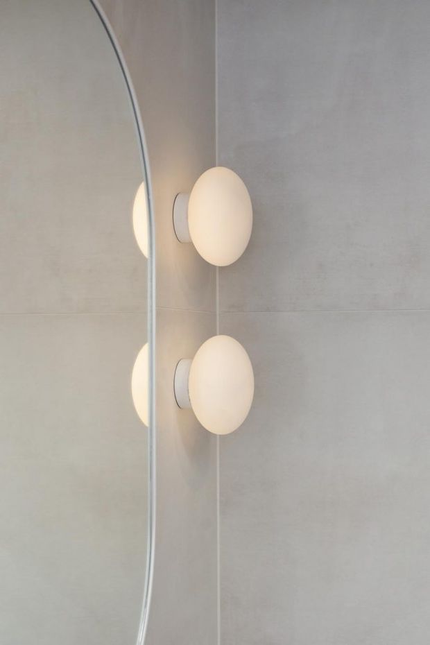 Close-up of two minimalist oval wall sconces with warm white diffused shades mounted on a pale grey tiled or plastered wall. A curved mirror at the left edge reflects the fixtures, creating repeated glowing forms. The composition is clean, understated, and contemporary.