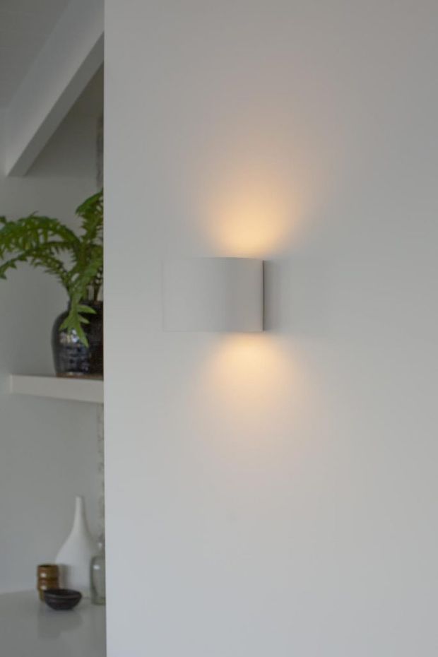 Close-up interior view of a minimalist white wall featuring a compact rectangular wall sconce casting warm up-and-down light. Built-in open shelving with a leafy plant and neutral decorative vessels is partially visible to the left.