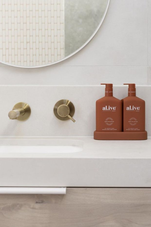 Close-up of a refined bathroom vanity with pale tiled walls, a large round mirror, brushed brass wall-mounted tapware, white integrated basin and benchtop, and warm terracotta-toned bathroom product bottles on a matching tray. Light timber cabinetry or flooring is visible below.