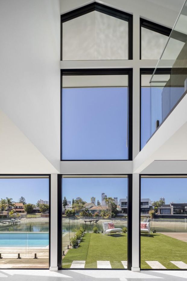 Bright contemporary interior overlooking a landscaped backyard and swimming pool. The space is defined by tall black-framed floor-to-ceiling glazing, a double-height void, and a glass balustrade at an upper-level stair or landing. A clear urban skyline is visible beyond the garden.