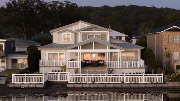 Front exterior view of a large light-coloured multi-level house with prominent covered verandas, white railings, gabled roof forms, and extensive waterfront-facing decking or fencing. The home sits in a suburban setting with wooded hills behind it and reflections visible in the water in the foreground.