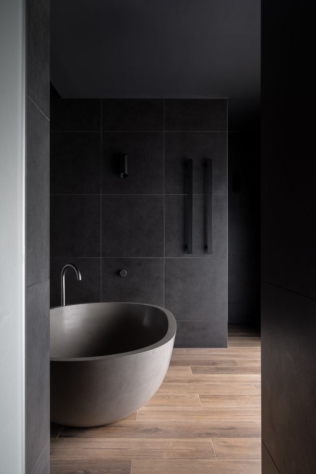 A moody, minimalist bathroom viewed through a doorway, featuring a freestanding oval bathtub, dark large-format tiled wall, black fixtures and cabinetry, and warm wood-look flooring. The restrained palette and concealed lighting create a refined, dramatic atmosphere.