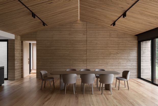 A warm, contemporary dining area with a long rectangular dining table and upholstered chairs, enclosed by textured earth-toned walls and extensive glazing. The room features a dramatic slatted timber ceiling following a pitched roof form, timber flooring, dark-framed windows and doors, and minimal linear lighting. An adjacent living area and hallway are visible at the edges.