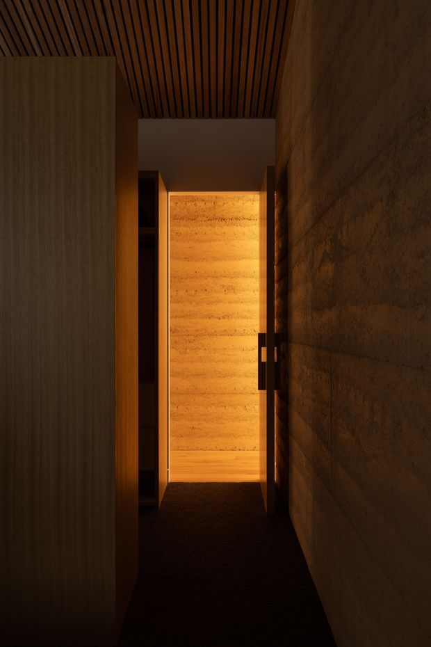 A narrow, dimly lit interior passage with a warm, atmospheric design. The corridor has dark carpet flooring, a timber slatted ceiling, a textured earthen or stone-like wall on the right, and dark timber cabinetry or wall panelling on the left. A warmly illuminated timber-lined opening or doorway draws the eye at the end of the passage.