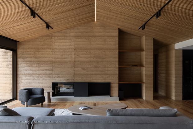 Contemporary living room with a low grey sectional sofa, dark lounge chair, rounded wood coffee tables, and a minimalist built-in media wall. Warm timber flooring and a richly textured wood-lined ceiling complement the earthy, horizontally striated wall finish. Black track lighting and dark-framed glazing add contrast, while open timber shelving and concealed cabinetry provide integrated storage.