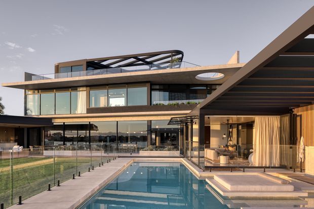 A contemporary multi-level residence arranged around a long rectangular swimming pool and landscaped courtyard. The home features broad floor-to-ceiling glazing, projecting flat roof planes, warm timber soffits and cladding, dark metal framing, extensive outdoor entertaining areas, and a rooftop terrace with a lightweight pergola structure. A covered outdoor lounge opens directly toward the pool.