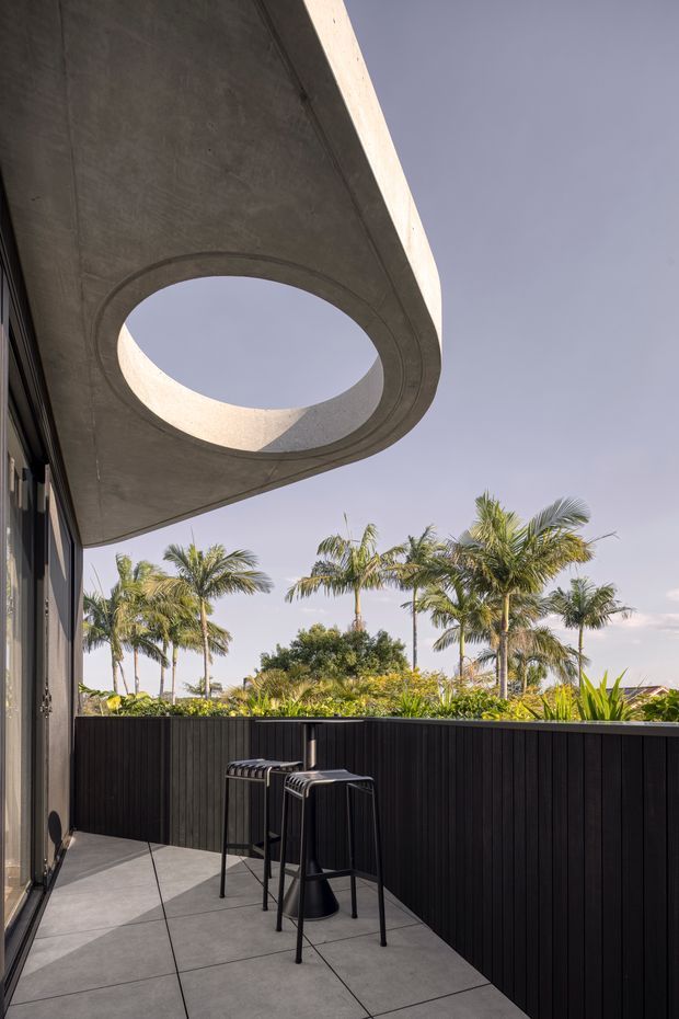 A contemporary covered outdoor balcony or terrace with a dramatic curved concrete soffit and large circular skylight opening. The terrace has a dark vertical-slat privacy balustrade, large grey tiled paving, and a small black bar-height table with stools. Tropical palms and dense planting are visible beyond the railing, creating a warm resort-like setting.