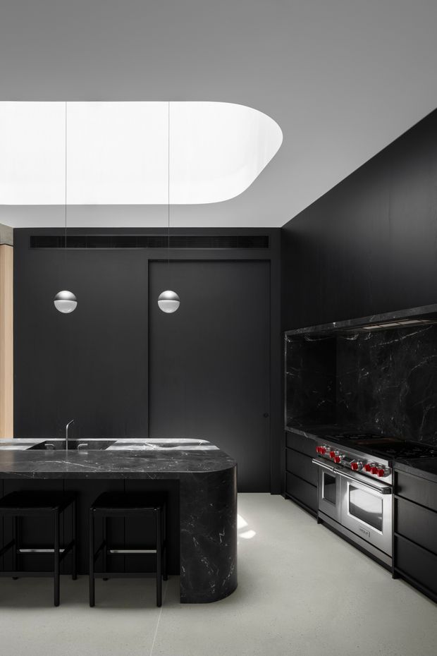 A dramatic contemporary kitchen dominated by matte black cabinetry and walls, with a dark veined stone island and matching splashback. The island includes a sink and seating for several people on black stools. A large stainless-steel range cooker with red controls is integrated into the cabinetry on the right. The room has a pale smooth floor, spherical wall lights, and a sculptural white suspended ceiling light, creating a high-contrast, moody interior.