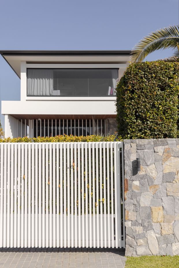 Street-facing view of a contemporary two-storey residence with a flat roof, crisp white rendered walls, expansive dark-framed glazing, vertical white privacy screens, and a tall white slatted gate. A rough stacked-stone boundary wall and manicured hedge frame the frontage, with paved access in the foreground and a palm visible at the side.