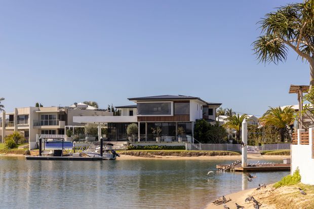 Exterior view across a calm canal toward a contemporary waterfront residence. The two-storey home has a low-pitched dark roof, white rendered walls, extensive glazing, and dark timber-toned facade panels. A private jetty and moored boat sit along the landscaped canal edge, with neighboring modern homes and palm-like planting visible in the background.