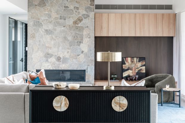A bright contemporary living room viewed from behind a black fluted console. The room features a large irregular natural-stone fireplace wall with a built-in horizontal firebox, warm timber wall cabinetry, a dark wood panelled media unit, light upholstered seating, and a brass-toned floor lamp. Glazing and pale curtains bring in abundant natural light.