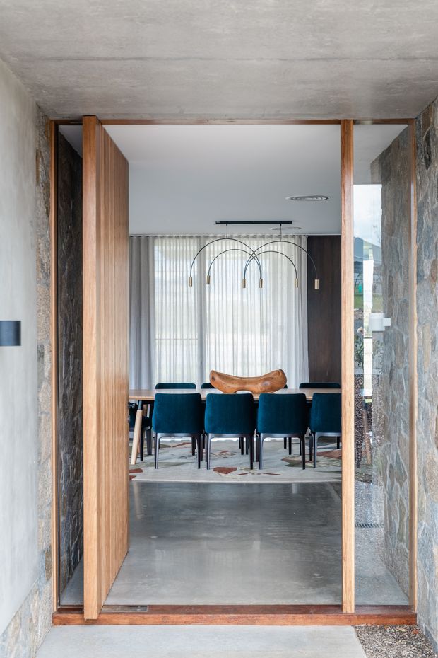 View through a tall timber-framed opening into a refined dining room. The room features a long dining table with deep teal upholstered chairs, a sculptural wooden centrepiece, a linear multi-arm pendant light, sheer curtains, dark timber wall panelling, a polished concrete floor, and textured stone walls surrounding the opening.