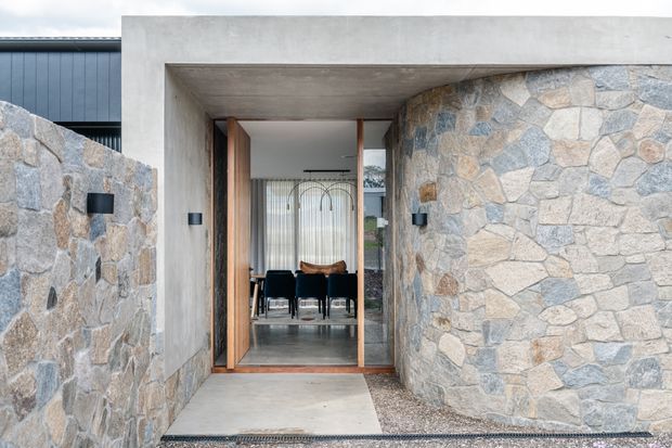 A contemporary residential entryway framed by a deep raw-concrete portal and curved walls clad in irregular grey and beige natural stone. Timber-framed glazed doors open toward a dining area, with minimalist black wall lights and a polished concrete entry path.
