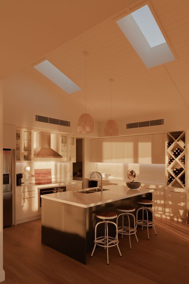 Warm, contemporary kitchen interior with a large central island, integrated sink and white cabinetry. The room features a timber-lined pitched ceiling with two roof skylights, pendant lights with woven shades, timber flooring, a stainless-steel rangehood, and a built-in wine display at the right. Strong low-angle sunlight creates a rich amber atmosphere.