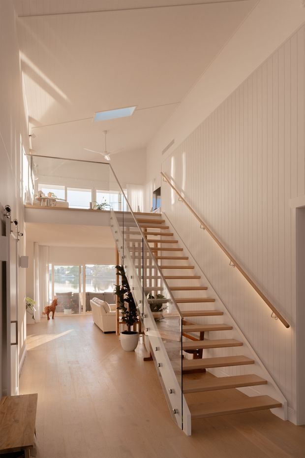 Bright, airy contemporary interior centred on an open timber staircase with glass balustrades and a timber handrail. The staircase rises through a double-height hallway or living area, with white vertical wall panelling, pale timber flooring, a skylight, ceiling fan, and large glazed openings. A partially visible living room and upper-level landing are in the background, with warm natural light throughout.
