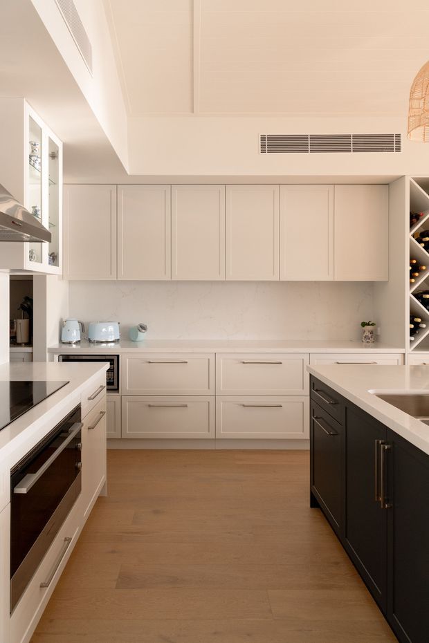 A bright, refined contemporary kitchen with a warm neutral palette. The room features a large central island with a dark charcoal cabinetry base and pale stone benchtop, extensive soft-white shaker-style cabinetry, integrated appliances, a pale stone splashback, timber flooring, a built-in wine rack, and understated pendant lighting. The layout appears spacious with a high ceiling and clean, uncluttered detailing.