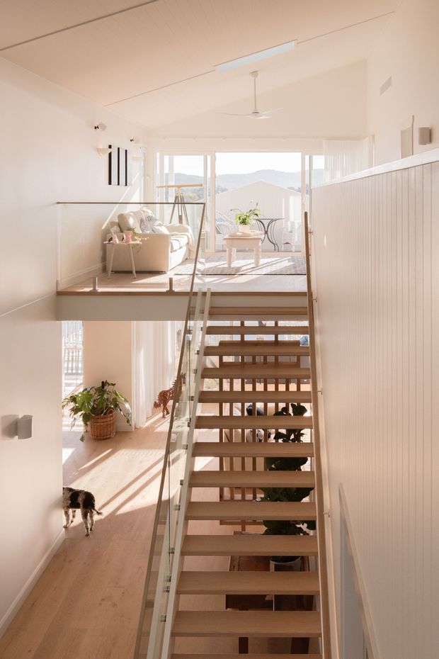 Bright, airy interior view looking up an open-riser timber staircase with a clear glass balustrade. The stair connects a lower hallway to an elevated living area furnished with a light sofa and coffee table, with large windows overlooking distant hills. White walls and vertical wall panelling, pale timber flooring and abundant natural light create a clean contemporary split-level home. A ceiling fan and linear ceiling light are visible.