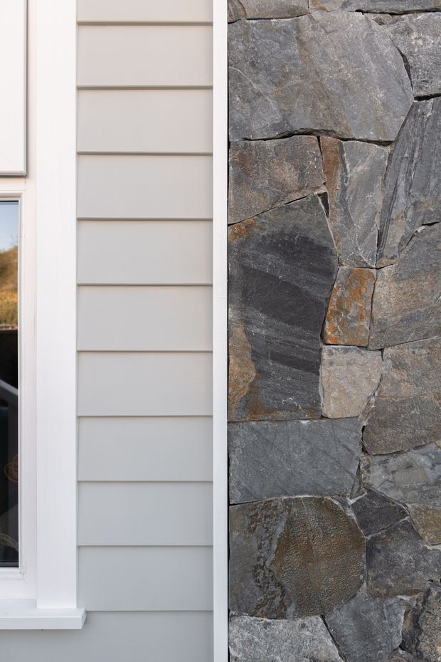 Close-up exterior detail showing pale grey horizontal weatherboard cladding beside a white-trimmed window and an adjacent wall of irregular dark grey natural stone masonry. The image emphasizes the contrast between smooth painted cladding and rugged stonework.