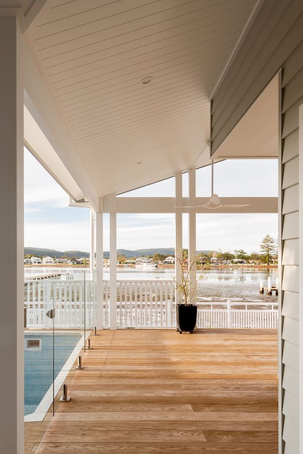 A bright covered outdoor deck overlooking a calm waterfront, with warm timber decking, white balustrades and posts, glass pool fencing, a partially visible swimming pool, and a white-lined ceiling. The setting includes a retractable awning, ceiling fan, and views across the water to low-density homes and tree-covered hills.