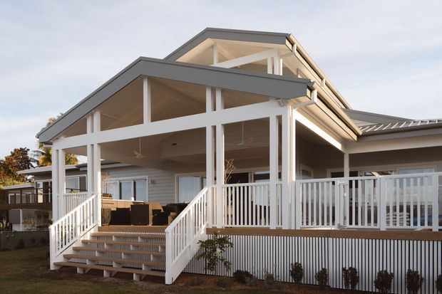Exterior view of a light-coloured, single-storey residential home with a prominent covered veranda, pitched gabled roof forms, white structural posts and balustrades, weatherboard-style cladding, and broad outdoor entertaining space accessed by stairs. The design has a relaxed traditional coastal character with open timber-framed porch structures.