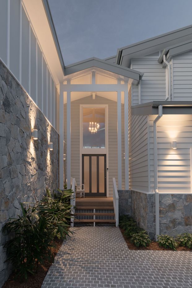 A narrow landscaped entry courtyard leading to a covered front entrance. The approach is paved in small grey brick pavers and bordered by a rugged natural-stone wall on one side and a light-grey weatherboard-clad house on the other. The entry has a pitched gabled canopy, tall glazed timber door, white trim, exterior lighting, and steps with dark metal balustrades. Warm interior and exterior lighting create an inviting dusk atmosphere.