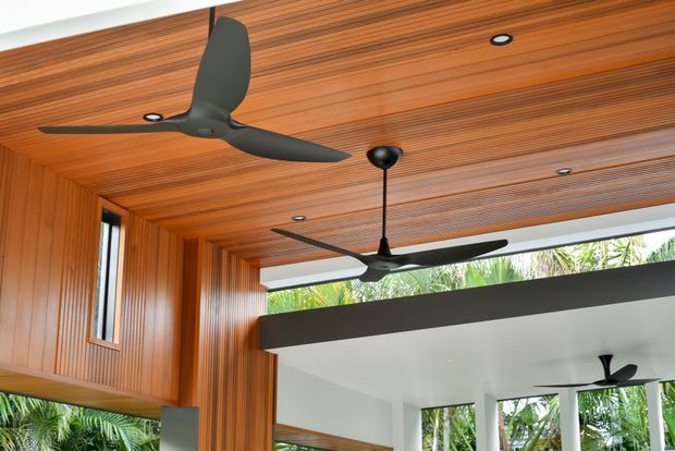 A contemporary interior featuring a richly stained timber-lined ceiling and walls, recessed downlights, and multiple matte-black ceiling fans. A large opening reveals a bright adjoining space with white surfaces, structural columns, and lush tropical planting beyond.