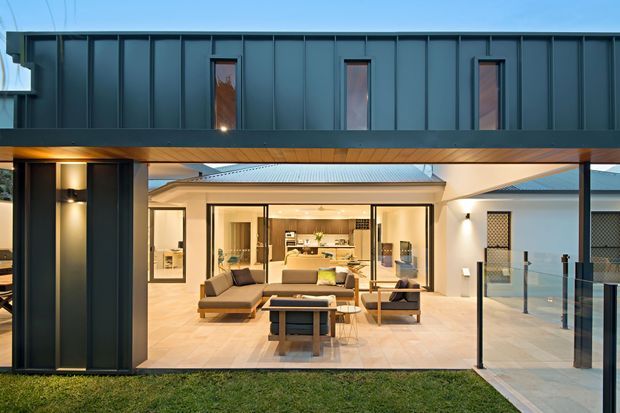 Exterior view of a contemporary multi-level home at dusk, with dark vertical metal cladding forming an upper volume and a broad covered outdoor living area below. The open glazing reveals a warm, modern living room with sofas and a central coffee table, while glass balustrades, pale paving, recessed lighting, and a light metal roof are visible.