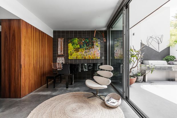 A refined compact interior lounge or entry area with a dramatic dark tiled feature wall, extensive warm timber built-in cabinetry, concrete flooring, a sculptural white lounge chair, and a large round natural-fibre rug. Full-height glazing opens onto a bright planted courtyard or terrace. A colourful artwork and a small desk or console occupy the rear of the space.