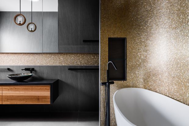 A sophisticated contemporary bathroom featuring a floating timber vanity with dark edging, a black countertop basin and wall-mounted tapware, dark grey cabinetry and wall panels, and a freestanding white bathtub. The walls are clad in a shimmering gold mosaic tile, creating a luxurious feature surface, with black-framed mirrors and pendant lighting adding contrast.