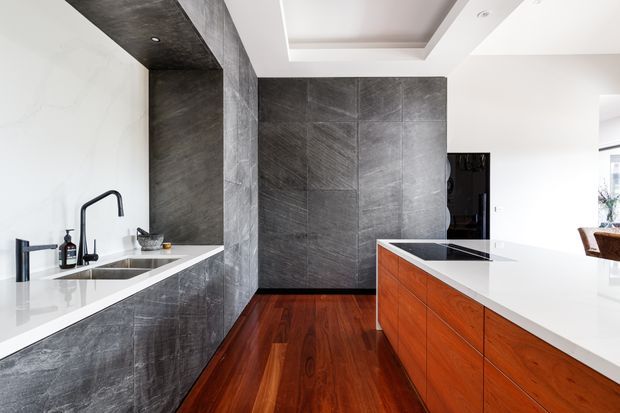 Contemporary kitchen featuring dark grey stone-look cabinetry and wall cladding, white waterfall-edge benchtops, warm timber floors, and a timber island with integrated cooktop. The space has a clean, refined finish with recessed ceiling detailing and strong contrast between dark surfaces, white counters, and reddish-brown wood.