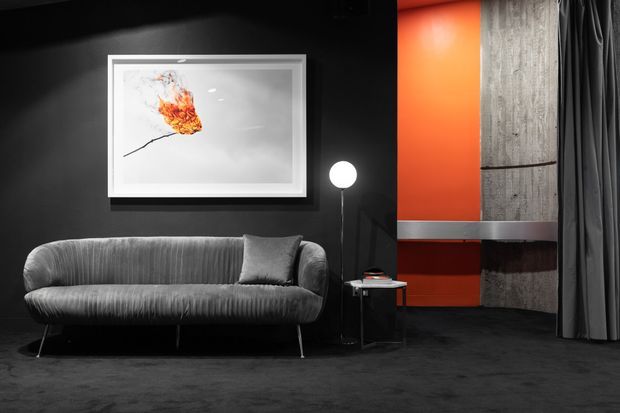 Moody, contemporary living room with a sculptural grey sofa against a matte black wall, a framed artwork, slim floor lamp, and small side table. A vivid orange feature wall and vertically textured concrete-like panel create a strong contrast, with dark curtains and flooring completing the dramatic monochrome palette.